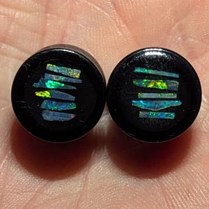 Könnte beinhalten: Paar schwarze Ohrdehner mit schimmerndem grünen und blauen Opal-Inlay. Das Inlay ist in einem geometrischen Muster aus horizontalen Linien.