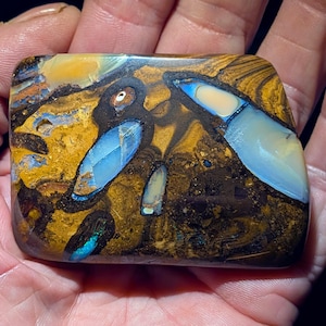 May include: A rectangular, polished opal stone with a unique pattern of blue, yellow, and brown hues. The stone features iridescent blue sections and a dark matrix, creating a striking visual contrast. The opal is held in a hand.