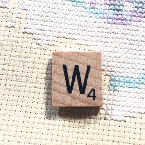 May include: A square wooden game tile featuring the black letter W and the number 4 printed on its surface, resting on a piece of cream-colored Aida fabric with a partially completed cross-stitch pattern in shades of purple and light blue.