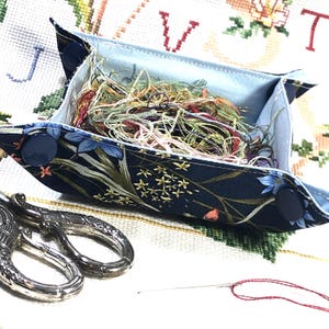 May include: A dark blue fabric tray with a floral pattern, holding colorful embroidery floss. Silver scissors and a needle with red thread are in the foreground. The tray is secured with black snaps.
