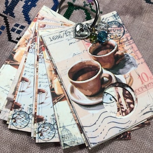 May include: A set of coffee-themed paper tags on a silver ring. The tags display images of coffee cups, stamps, and vintage text. A heart-shaped charm and beads hang from the ring. The colour scheme includes browns, creams, and beige.