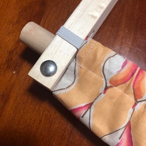 May include: A close-up shot of a wooden frame with a fabric sleeve. The fabric features a pattern of orange, pink, and brown shapes. A grey elastic band secures the fabric. A metal rivet is visible.