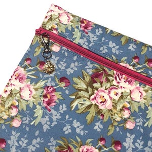May include: A fabric pouch featuring a blue background with a floral pattern of pink and white peonies and green leaves. The pouch includes a bright pink zip closure and a silver-toned metal flower charm attached to the zip pull.