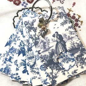 May include: A set of blue and white patterned paper tags held together by a silver ring. The tags feature a classic toile design with scenes of couples and landscapes. A decorative charm with beads and a leaf hangs from the ring.