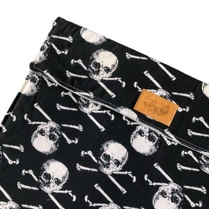 May include: A black fabric item featuring a repeating white skull and crossbones pattern. The fabric has a folded top edge and a rectangular tan leather-like patch sewn onto the front with the text Simply Needle embossed in a cursive script.