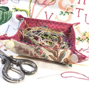 May include: A patterned fabric tray with a pink interior contains a colourful collection of embroidery floss. Silver scissors and a needle with red thread are placed nearby, indicating a craft project.