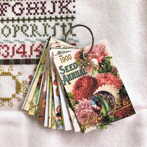 Peut inclure: Un ensemble de cartes de paquets de graines vintage sur un anneau argenté. Les cartes présentent des illustrations florales colorées et du texte, dont "1900 Seed Annual" et "D.M. Ferry & Co. Detroit Mich.". Un charme avec une fleur et une perle est suspendu à l'anneau.