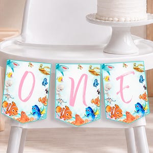 May include: A white high chair with a cake on a stand and a three-piece banner hanging from the tray. The banner features watercolor illustrations of sea creatures and the letters O, N, and E in a large, light pink script font on each panel.