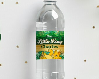 Puede incluir: Una botella de agua de plástico transparente con tapa blanca. La etiqueta dice "Our Little King Charles" con una ilustración de león de dibujos animados. La botella está rodeada de hojas decorativas y pequeñas estrellas doradas.