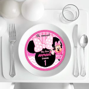 May include: A white plate with a Minnie Mouse design. The plate has a pink and black colour scheme with the text "Oh, Toodles!" and "Our little Armani turns 1". A fork, knife, and spoon are on the table.
