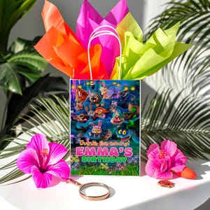 A birthday bag with a colorful design featuring cartoon characters, placed on a table with a pink hibiscus flower, a magnifying glass, and a pink hibiscus.