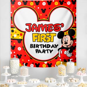 May include: A red and white Mickey Mouse-themed banner with the text "JAMES' FIRST BIRTHDAY PARTY." The banner features Mickey Mouse and decorative elements. Various candies are displayed on white cake stands and in glass jars.