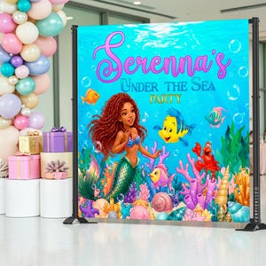 May include: A vibrant backdrop for a party, featuring a mermaid with red hair, a blue top, and a green tail. The backdrop has the text "Serenna's Under The Sea Party" with cartoon fish, coral, and bubbles. The backdrop is supported by black metal stands.