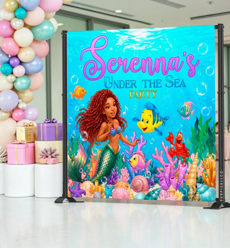 May include: A vibrant backdrop for a party, featuring a mermaid with red hair, a blue top, and a green tail. The backdrop has the text "Serenna's Under The Sea Party" with cartoon fish, coral, and bubbles. The backdrop is supported by black metal stands.