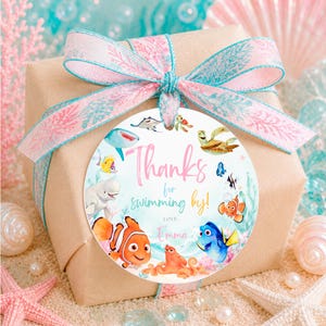 May include: A square gift box wrapped in brown paper with a pink and teal coral-patterned ribbon. A round tag features colorful cartoon sea creatures and the text "Thanks for swimming by! LOVE, Emma" printed in pink and teal script and sans-serif fonts.