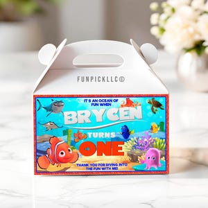 Peut inclure: Boîte blanche à pignon avec un design coloré sur le thème de l'océan. La boîte présente des poissons de dessins animés, un requin et le texte "Brycen Turns One". La boîte comprend le texte "Thank you for diving into the fun with me!"