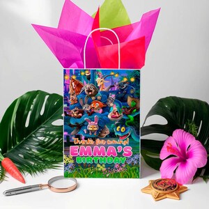 A colorful birthday bag with a cartoon character design, surrounded by tropical leaves, a magnifying glass, and a pink hibiscus flower.