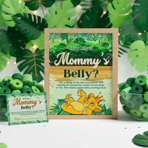 Puede incluir: Un letrero enmarcado para un juego de baby shower con el texto "How Big is Mommy's Belly?" y un león de dibujos animados. Hojas de papel verde y rollos de cinta verde rodean el letrero. También se ve una tarjeta más pequeña con el mismo texto.