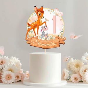 Puede incluir: Un adorno para tarta con un diseño de Bambi y Tambor, con el número 1. El adorno incluye el nombre "Samantha" en una pancarta de madera, rodeado de flores y mariposas. El adorno está colocado encima de una tarta blanca.
