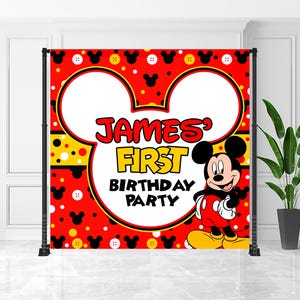 May include: A red backdrop with Mickey Mouse ears and a cartoon image of Mickey Mouse. The backdrop has the text "James' First Birthday Party" in red and yellow. The backdrop is supported by a black metal frame.