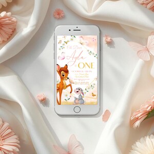 Op de afbeelding: Een smartphone toont een aangepaste uitnodiging met de tekst "Oh Deer, Ayla is ONE." Het ontwerp bevat een cartoon hert en konijn, vlinders en bloemendetails in goud, roze en wit. De telefoon is omgeven door roze bloemen en vlinders.