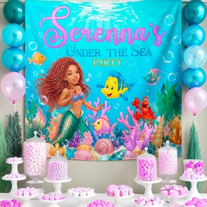 May include: A party backdrop with the text "Serenna's Under The Sea Party" featuring a mermaid and sea creatures. Balloons in teal and pink flank the backdrop. Tables display various pink candies and treats, creating a festive underwater theme.