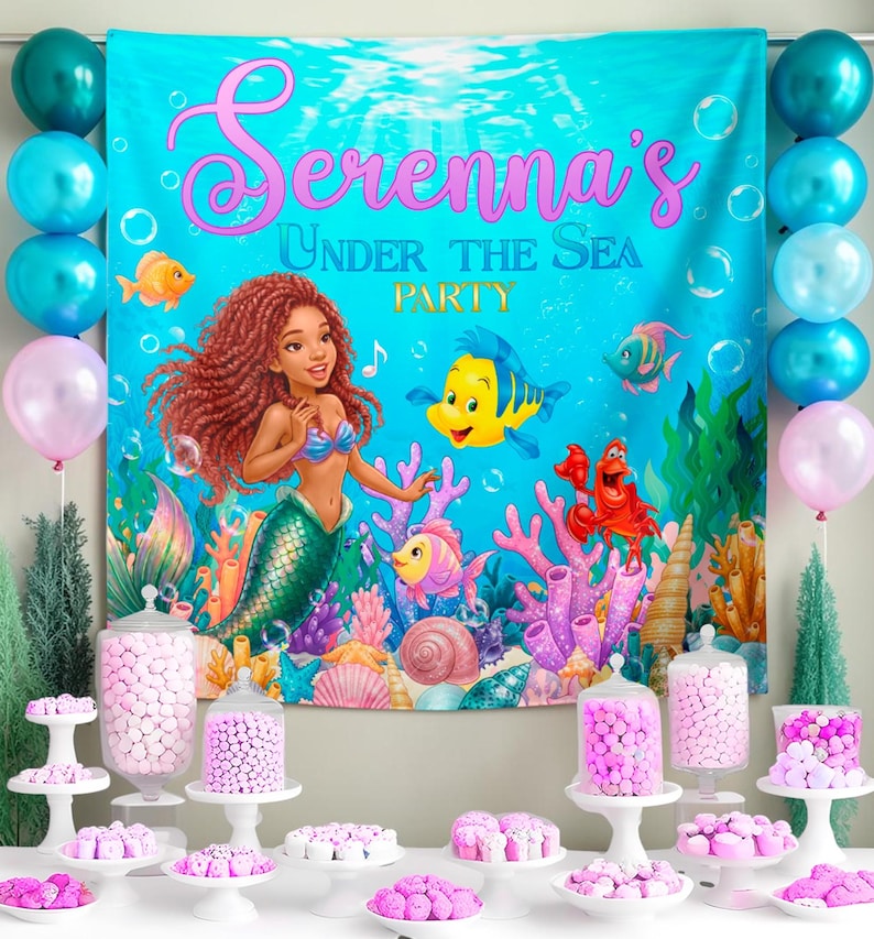 May include: A party backdrop with the text "Serenna's Under The Sea Party" featuring a mermaid and sea creatures. Balloons in teal and pink flank the backdrop. Tables display various pink candies and treats, creating a festive underwater theme.