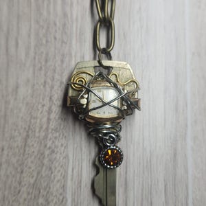 May include: A pendant featuring a vintage key wrapped in silver and gold-toned wire, centered with a small square watch face. The piece includes a dangling amber-colored circular gemstone and is attached to a thick, antique brass-colored chain.