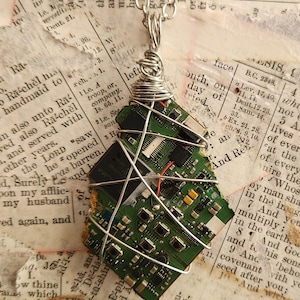 May include: A pendant made from a fragment of a green circuit board, wrapped in silver-toned wire and suspended from a matching silver-toned metal chain. The circuit board features visible electronic components, including small black chips.