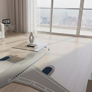 May include: A unique coffee table with a glass top, resting on a gray airplane-shaped rug. The rug features blue accents on the wings. A modern living room with a city view is in the background.