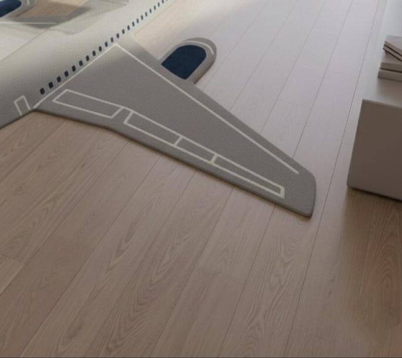May include: A gray airplane-shaped rug with white outline details and a dark blue accent. The rug is placed on a light-colored wooden floor. The design resembles an airplane wing and tail.