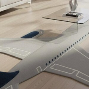 May include: A gray airplane-shaped rug with white outline details and dark blue accents. The rug is placed on a light wood floor, with a glass coffee table and decor in the background.
