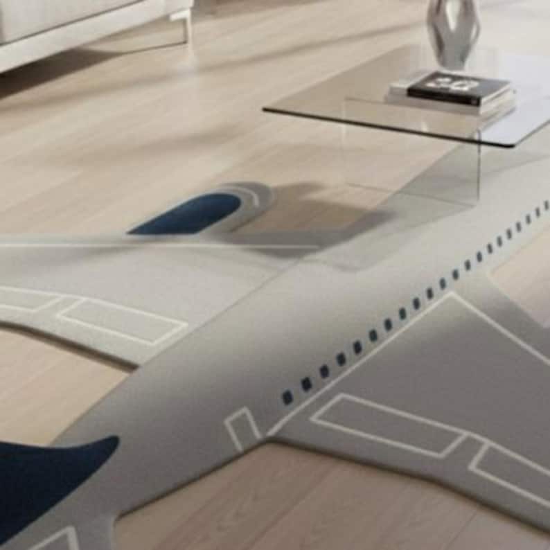 May include: A gray airplane-shaped rug with white outline details and dark blue accents. The rug is placed on a light wood floor, with a glass coffee table and decor in the background.