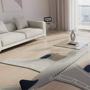 May include: A gray airplane-shaped rug with white detailing and navy blue accents. The rug is placed on a light wood floor in a modern living room setting. A glass coffee table and a white sofa are also visible.