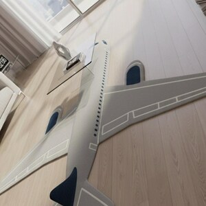 May include: A gray airplane-shaped rug with white trim and dark blue accents. The rug is placed on a light wood floor. A glass table and a decorative object are visible in the background.