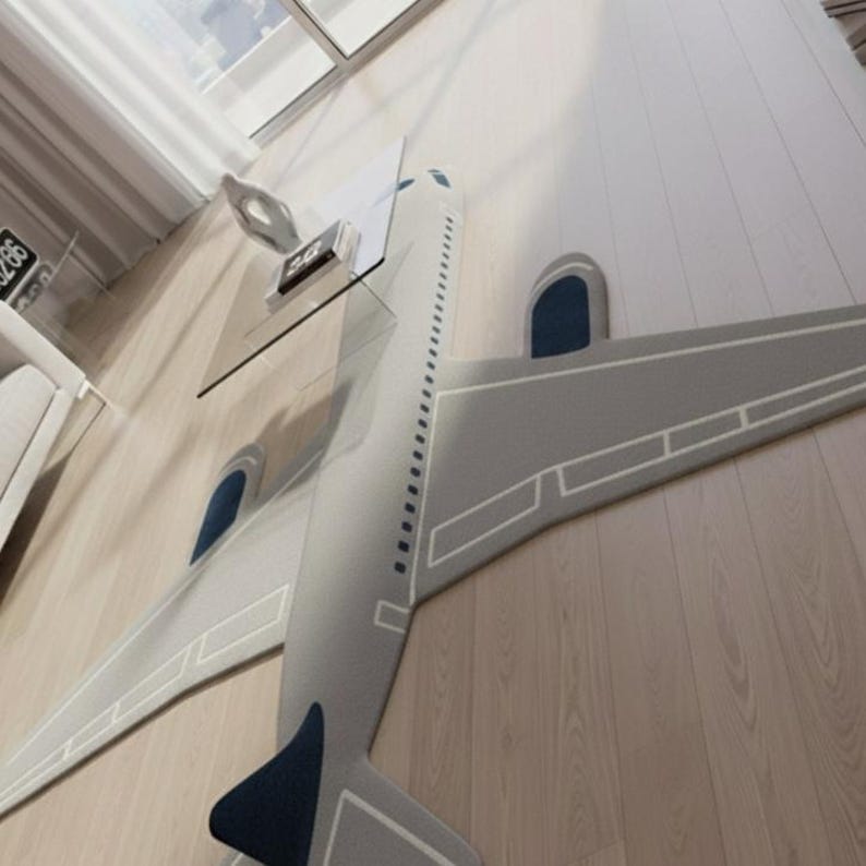 May include: A gray airplane-shaped rug with white trim and dark blue accents. The rug is placed on a light wood floor. A glass table and a decorative object are visible in the background.