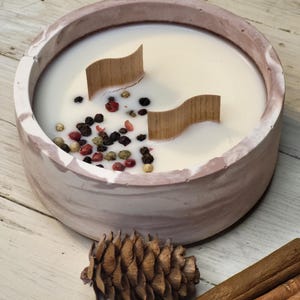 May include: A round, marbled pink and white candle in a concrete bowl. The candle has two wooden wicks and is decorated with peppercorns. A pine cone and cinnamon sticks are next to the candle.