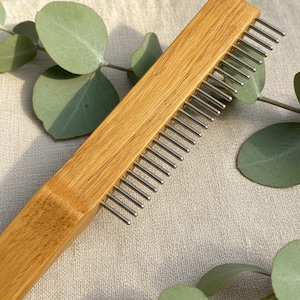 May include: A wooden comb with metal tines. The comb is made of light brown wood and has a rectangular shape. The metal tines are evenly spaced and extend from the comb's body. The comb is set against a neutral background with green leaves.