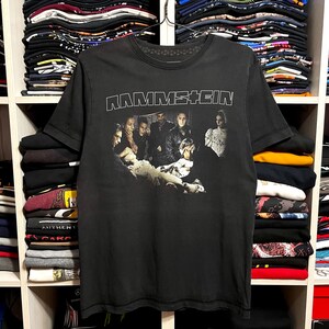 May include: Black Rammstein band t-shirt featuring the band's name in white block letters across the top. Below the text is a black and white image of the band members. The shirt is hanging on a metal hanger.