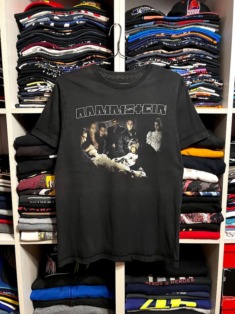 May include: Black Rammstein band t-shirt featuring the band's name in white block letters across the top. Below the text is a black and white image of the band members. The shirt is hanging on a metal hanger.