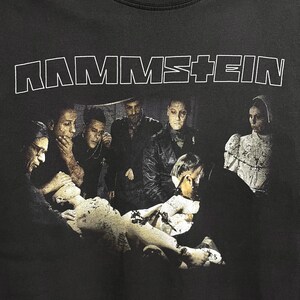 May include: Black t-shirt featuring the band name "RAMMSTEIN" in white block letters across the top. The shirt displays a dark, artistic image of several figures surrounding a statue.