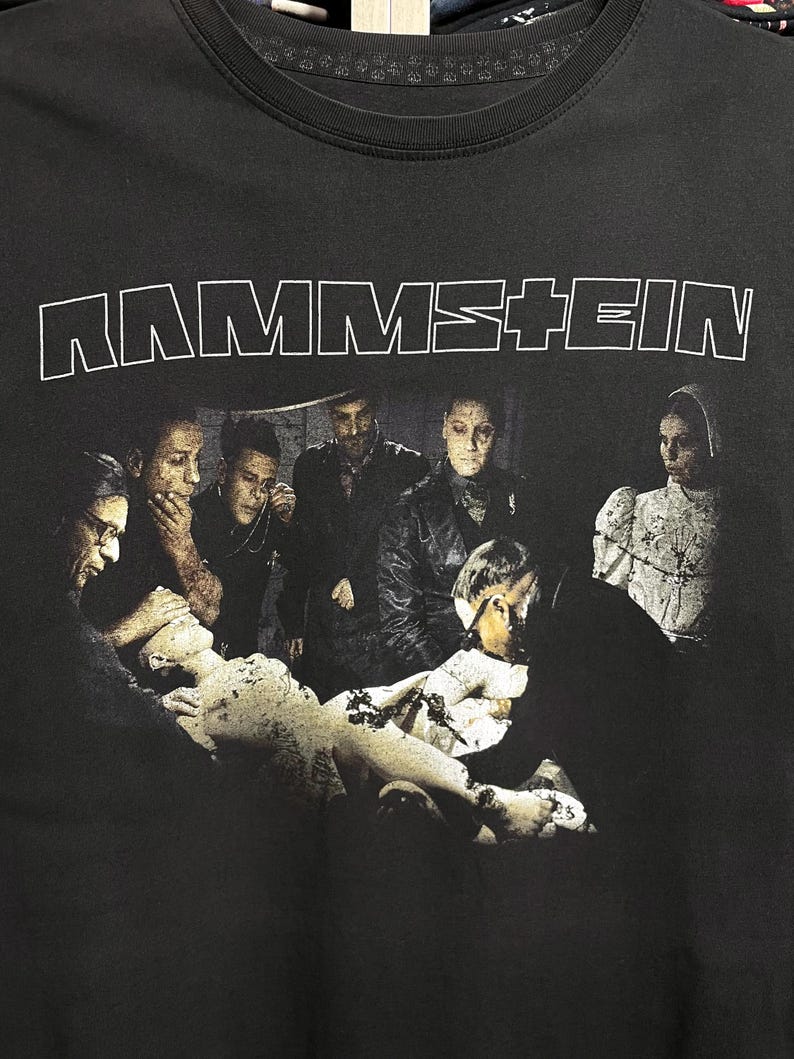 May include: Black t-shirt featuring the band name "RAMMSTEIN" in white block letters across the top. The shirt displays a dark, artistic image of several figures surrounding a statue.