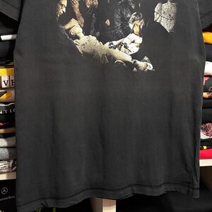 May include: Black t-shirt featuring the band name "RAMMSTEIN" in white block letters at the top. Below, a grayscale image depicts a group of people in a dark, gothic setting. The shirt is made of a soft, dark fabric.