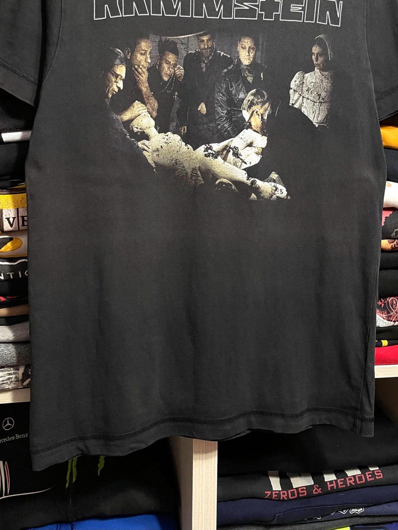May include: Black t-shirt featuring the band name "RAMMSTEIN" in white block letters at the top. Below, a grayscale image depicts a group of people in a dark, gothic setting. The shirt is made of a soft, dark fabric.