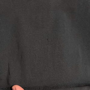 May include: Close-up of a black fabric with a visible texture. The fabric has a smooth appearance with a stitched hem at the bottom. The image shows a small dark spot on the fabric.