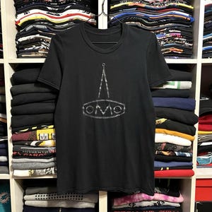 Könnte beinhalten: Ein schwarzes Kurzarm-T-Shirt mit einem weißen Grafikdruck auf der Brust. Das Design besteht aus einer nach oben zeigenden Dreiecksform über einem Oval, das die Buchstaben OMO enthält, alles in einem abgenutzten weißen Druckstil gehalten.