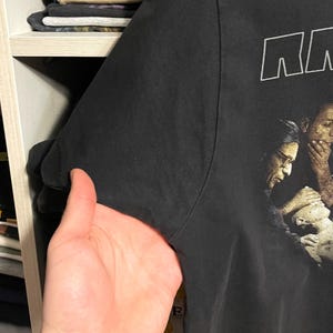 May include: Black t-shirt with a graphic print of a group of people. The word "NN" is printed in white block letters at the top. The shirt is held up by a hand, showing the sleeve and part of the shirt's side.