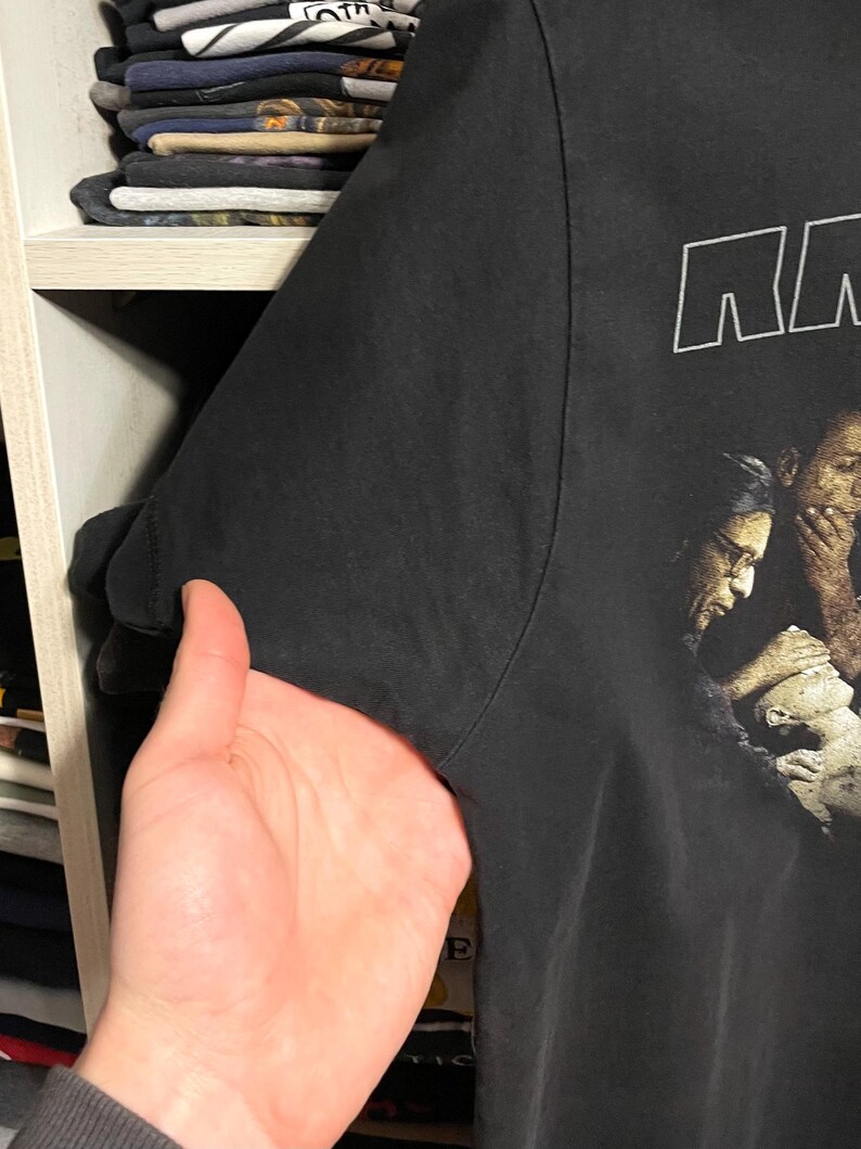 May include: Black t-shirt with a graphic print of a group of people. The word "NN" is printed in white block letters at the top. The shirt is held up by a hand, showing the sleeve and part of the shirt's side.