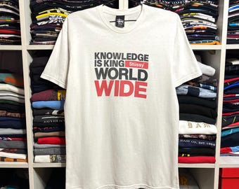 May include: A white t-shirt with the text "KNOWLEDGE IS KING WORLD WIDE" in black and red. The brand name "Stüssy" is above the words "WORLD WIDE". The shirt is hanging on a black hanger in front of a shelf filled with folded clothing.