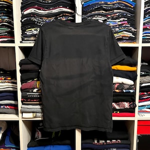 May include: A black t-shirt hangs in front of a shelf filled with folded clothing and hats. The t-shirt is plain and appears to be made of a soft material. The shelf is divided into compartments, each holding stacks of various items.